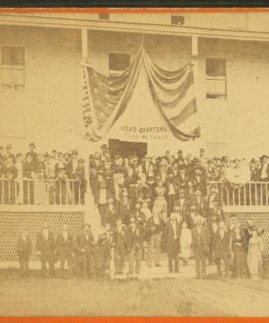 [Group of men and women posing on steps of large building, flag draped overhead, and a sign reading "Headquarters First ME Cavalry".] 1868?-1908