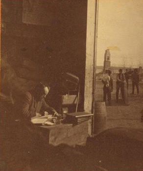 [A man working at a desk in a barn or shed with the ruins of Kineo house, after a fire, in the background.] 1870?-1880?