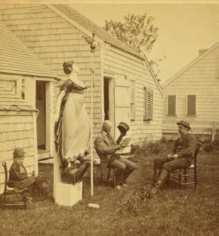 [View of men and a child in a yard with a wood carving of a woman holding a pole topped with a weathervane.] 1867?-1890?