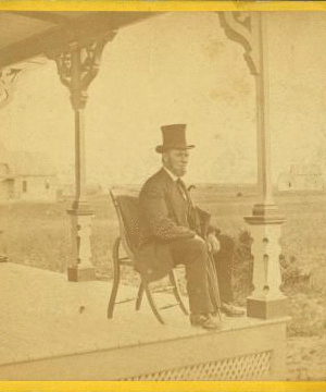 [View of a man with a beard and top hat on a porch.] 1868?-1880?