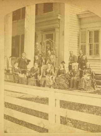 [Multi-generation male and female family members, wearing suits and dark dresses, pose before white-columned porch and trellis of multi-story clapboard house; white board fence in foreground.] 1860?-1915?