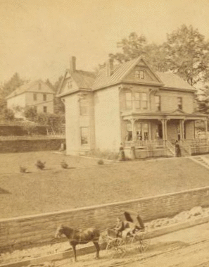 [Three-story frame house on terraced yard; people pose on porch; young man holds reins in one-horse carriage. Retaining wall and street under construction.] 1860?-1900?