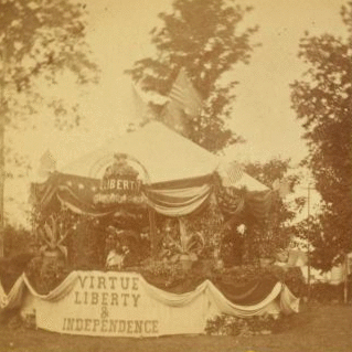 [Decorated tent [Fourth of July?], with American flags, ornamental plants, liberty bell in flowers, and drapery bearing phrase 'Virtue Liberty & Independence;' bearded, hatted man stands inside.] 1860?-1915?