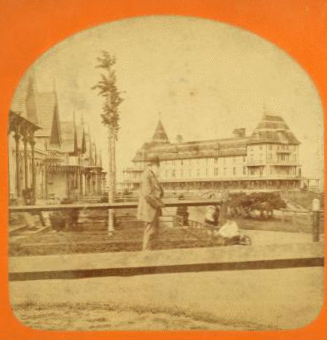 [View of cottages with a man standing, holding the handle of a toy wagon with a boy in it, Sea View House in the background.] 1865?-1885?