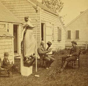 [View of men and a child in a yard with a wood carving of a woman holding a pole topped with a weathervane.] 1867?-1890?