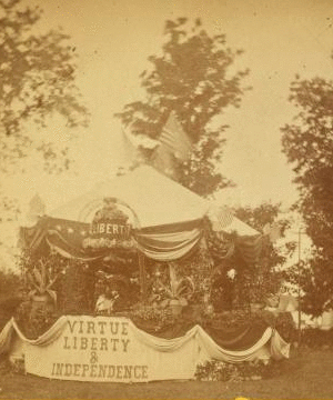 [Decorated tent [Fourth of July?], with American flags, ornamental plants, liberty bell in flowers, and drapery bearing phrase 'Virtue Liberty & Independence;' bearded, hatted man stands inside.] 1860?-1915?