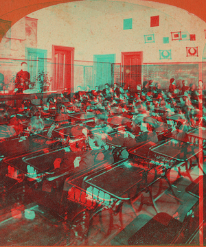 Woman teaches classroom, while the students in the right background write on the chalkboard