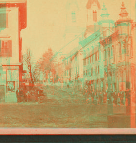 [View of people standing on a commercial street, church visible in background.] 1860?-1890?