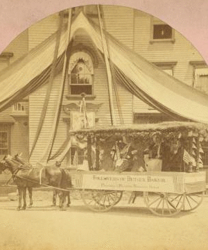 [Decorated wagon with women riding in it, banner on side reads "followers of Betsy Baker preparing and braiding domestic straw," in front of building with bunting.] 1865?-1880?