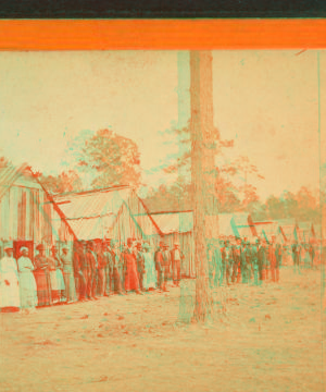 [Group lined up in front of a row of shacks.] 1868?-1900?