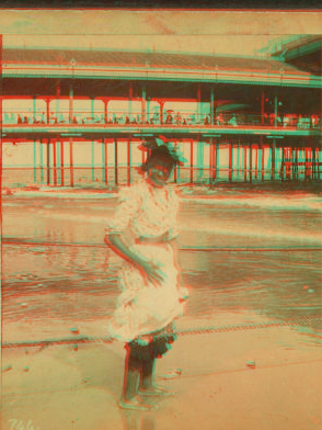[Young woman wading at the beach, in front of a covered pier.] 1868?-1900?